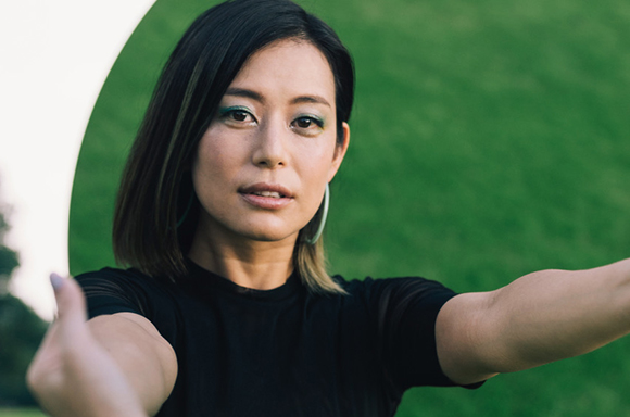 A woman with dark hair and green eyeshadow extends her arms toward the camera against a green background.