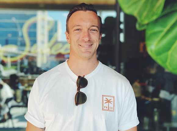 A smiling man wearing a white t-shirt with sunglasses tucked into the neckline.
