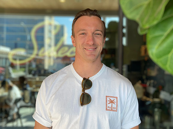 A smiling man wearing a white t-shirt with sunglasses hanging from the neckline.