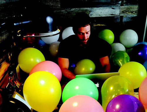 A man sits amidst a large collection of bright, multicolored balloons in a dark room.