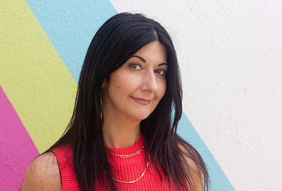 A woman with long dark hair and a red top smiles against a background of pastel-colored diagonal stripes.