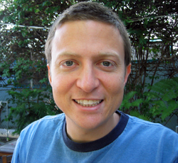 A smiling man with short brown hair wearing a blue crewneck shirt outdoors.