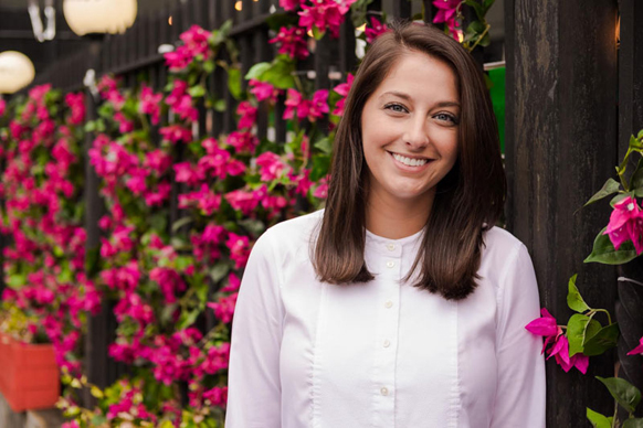 A smiling woman with long brown hair wearing a white button-down shirt, standing before bright pink flowers.