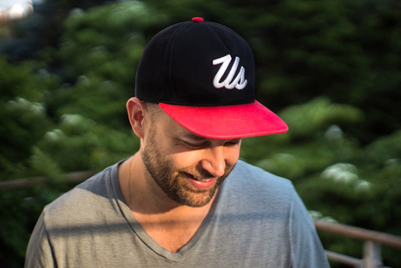 A man with a beard wearing a black baseball cap with a white 'Us' logo and a red brim.