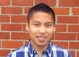 A young man with short dark hair smiling in a blue plaid shirt against a red brick wall.