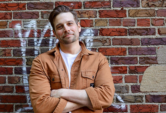 A man with short brown hair and a light beard wearing a brown button-down shirt over a white t-shirt.