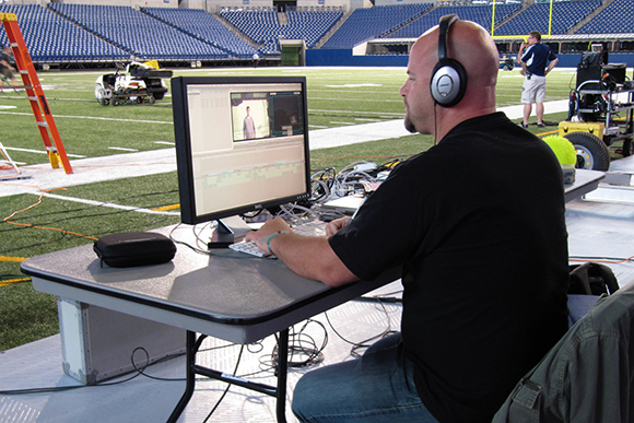 A man in a black shirt sits at a desk using a computer in a large sports stadium.