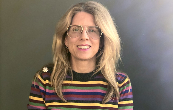 A smiling woman with long blonde hair and large glasses wearing a multi-colored striped sweater.
