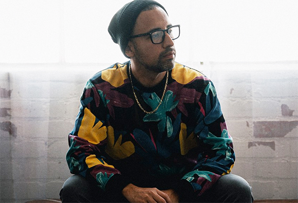 A man wearing a black beanie, glasses, and a colorful floral patterned sweatshirt sits against a brick wall.