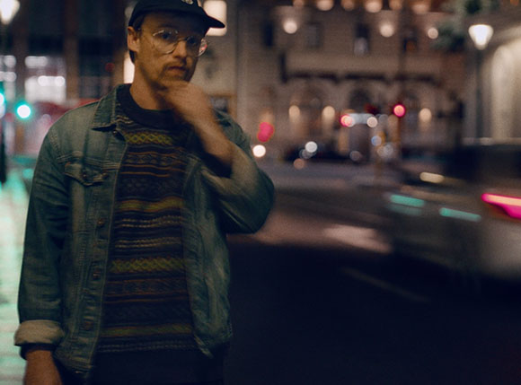 A man wearing glasses and a denim jacket over a patterned sweater stands in a city at night.