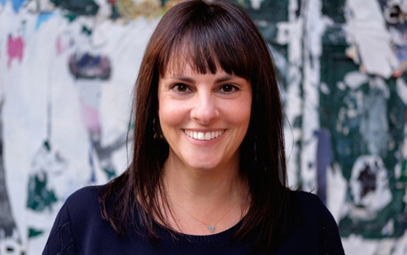 A smiling woman with dark hair and bangs wearing a dark blue top against a colorful, textured background.