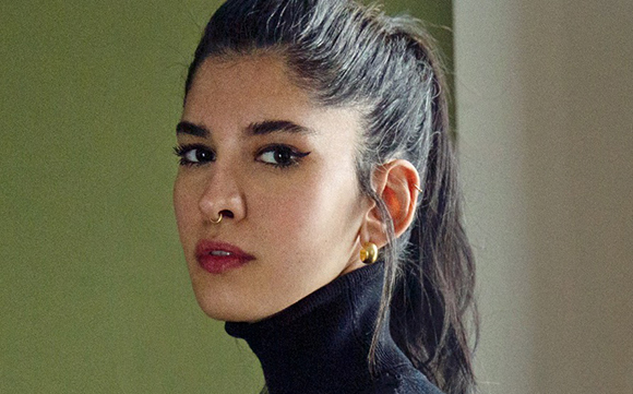 A woman with dark hair in a ponytail, wearing a black turtleneck and gold hoop earrings.