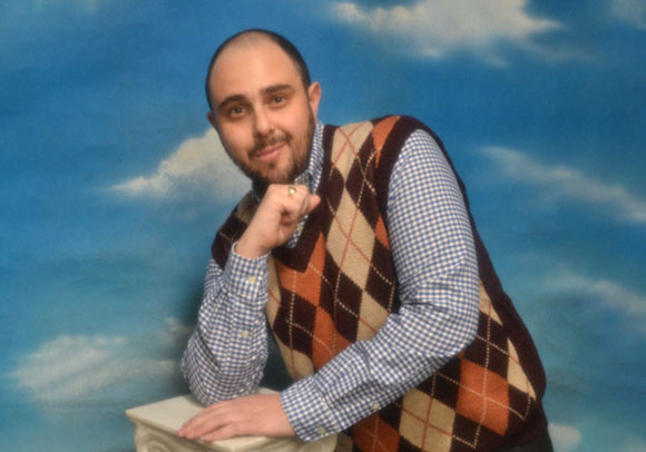 A man with a beard wearing a checkered shirt and an argyle sweater vest, posing against a blue sky background.