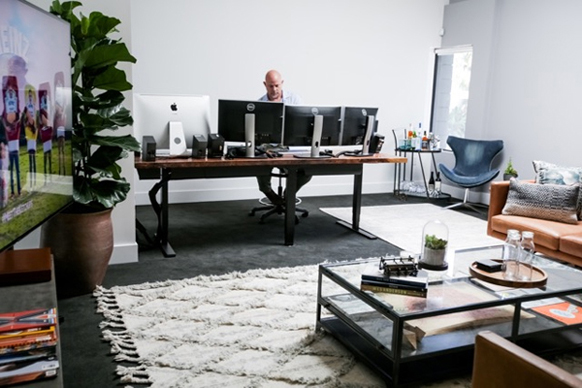 A man sits at a long wooden desk with three computer monitors in a bright, modern office.