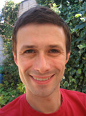 A smiling man with short brown hair wearing a red t-shirt in front of green foliage.