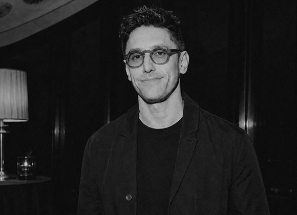 A man with dark hair and glasses wearing a dark blazer and t-shirt.