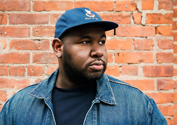 A man with a beard wearing a blue baseball cap and denim jacket stands in front of a brick wall.