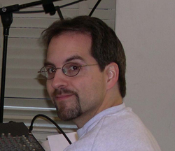 A man with short dark hair, glasses, and a goatee looking toward the camera in an indoor setting.