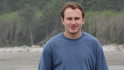 A man with short brown hair wearing a blue crewneck t-shirt, standing in front of a blurred forest background.