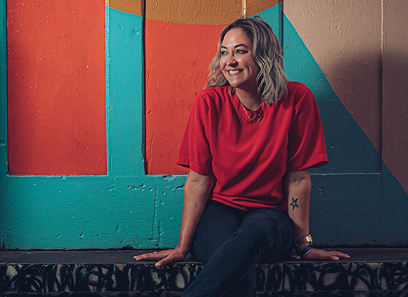 A smiling woman with blonde hair wearing a red t-shirt sits before a teal and orange geometric mural.