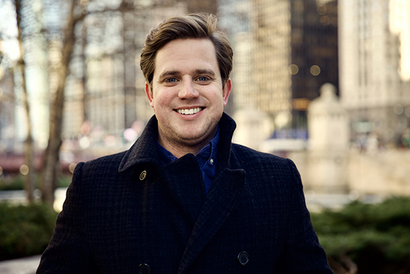 A smiling man with light hair wearing a dark navy pea coat in a city setting.