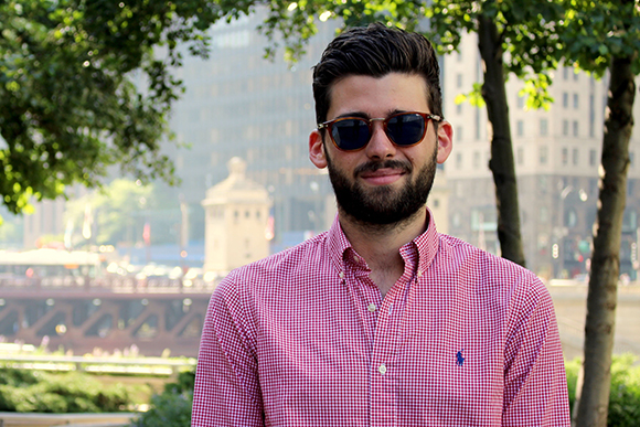 A bearded man wearing sunglasses and a red checkered shirt smiles in front of a city background.
