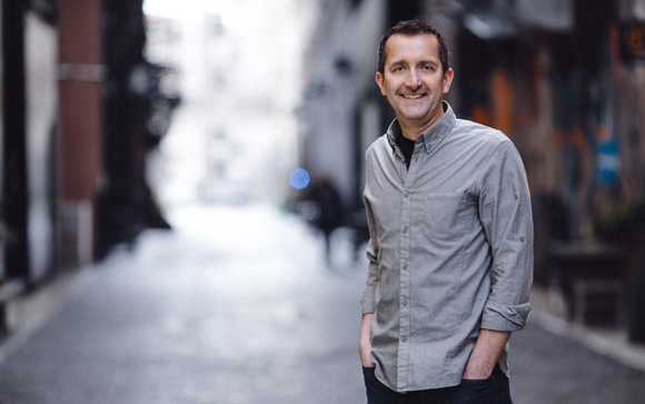 A smiling man in a grey button-down shirt stands in a blurred urban alleyway.