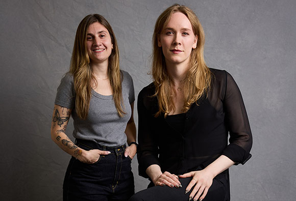 Two women with long hair posing together against a textured gray backdrop.
