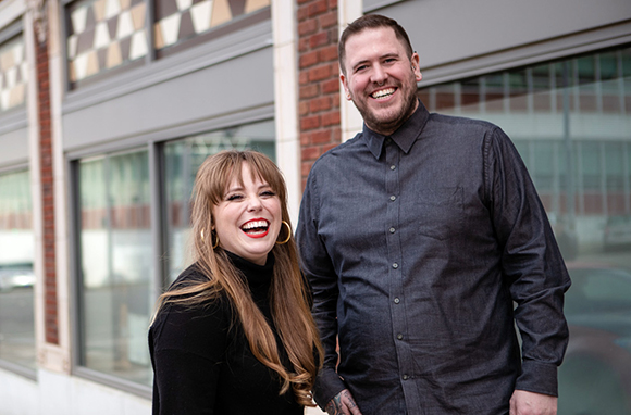 A smiling man in a dark button-down shirt and a woman with long hair and red lipstick standing together.