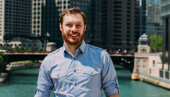 A smiling man with a beard wearing a light blue button-down shirt, standing before a city river and buildings.