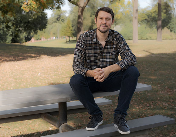A man in a plaid shirt and dark jeans sits on a grey picnic table in a sunlit park.