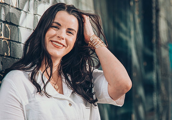 A smiling woman with long dark hair and gold bangles leans against a brick wall.