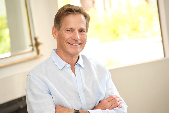 A middle-aged man with brown hair smiling and crossing his arms while wearing a light blue button-down shirt.