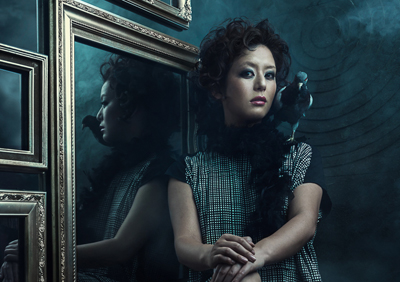 A woman with dark curly hair looks forward while standing beside a large gold-framed mirror.