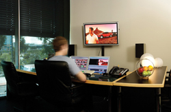 A person sits at a desk with two computer monitors and a large wall-mounted screen showing a video.