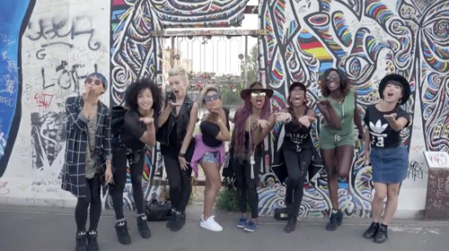 A diverse group of women posing and making expressive gestures in front of a colorful graffiti wall.