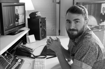 A bearded man sits at a desk with a computer, monitors, a telephone, and speakers in a black and white photo.