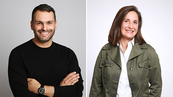 A man in a black sweater and a woman in an olive jacket smiling against a white background.