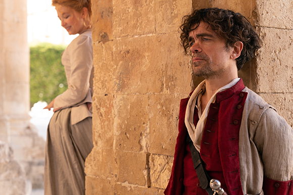 A man with dark curly hair in a red vest leans against a stone pillar, looking away from a woman in the background.