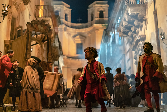 People in 18th-century clothing walk through a narrow, dimly lit European street at night.