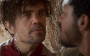 A man with curly dark hair and stubble stares intensely at another man in profile.