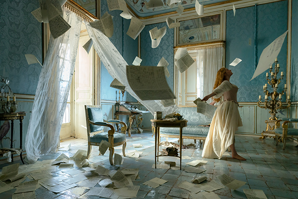 A woman in a long white dress stands in a blue ornate room as many sheets of paper fly through the air.