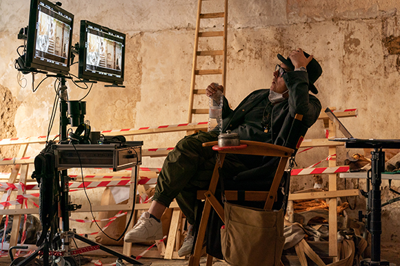 A person in a dark jacket and hat sits in a director's chair next to two film monitors in a rustic studio.
