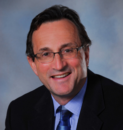 A man with dark hair and glasses wearing a dark suit and blue tie, smiling against a blue background.
