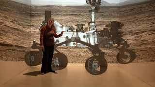 A woman in a red shirt stands before a large-scale mural of a planetary rover on a desert landscape.