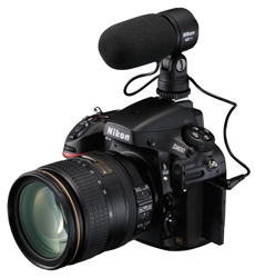 A black Nikon DSLR camera equipped with a large lens and an external shotgun microphone on top.