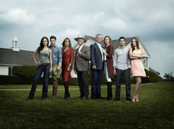 Seven people posing together in a line on a grassy field in front of a house under a cloudy sky.