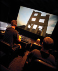 Two people sit in a dark theater watching a large screen displaying a ruined stone structure.