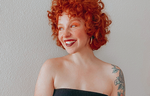 A smiling woman with curly red hair and a shoulder tattoo looking to the side.