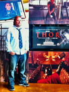 A man in a light blue shirt and tie stands beside posters for Thor and other films.